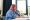 A plus size businesswoman with pink hair, wearing a blue shirt, sits at a desk looking thoughtfully off to the side.