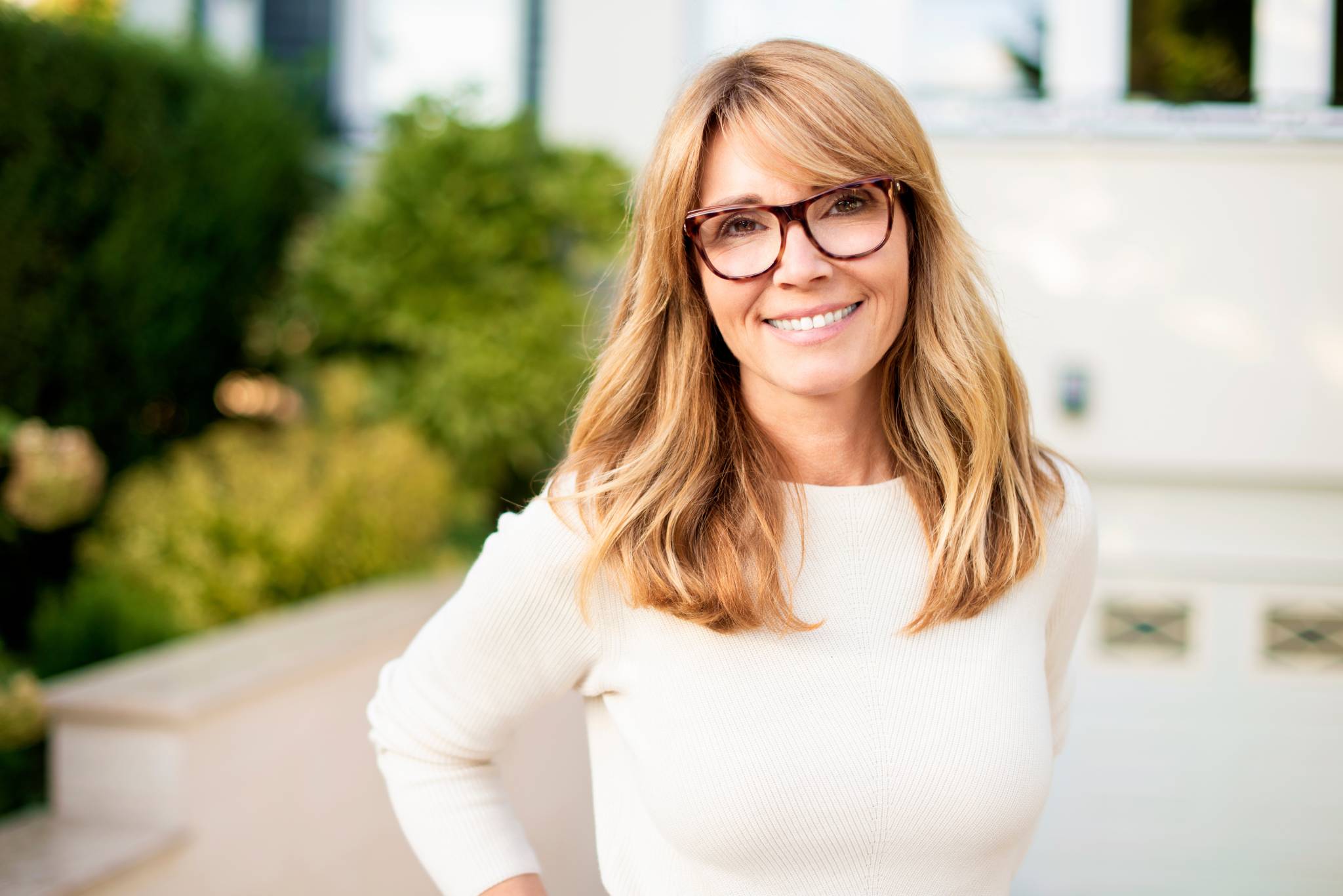 woman with blonde hair and glasses wearing a cream sweater, smiling outdoors with greenery in the background